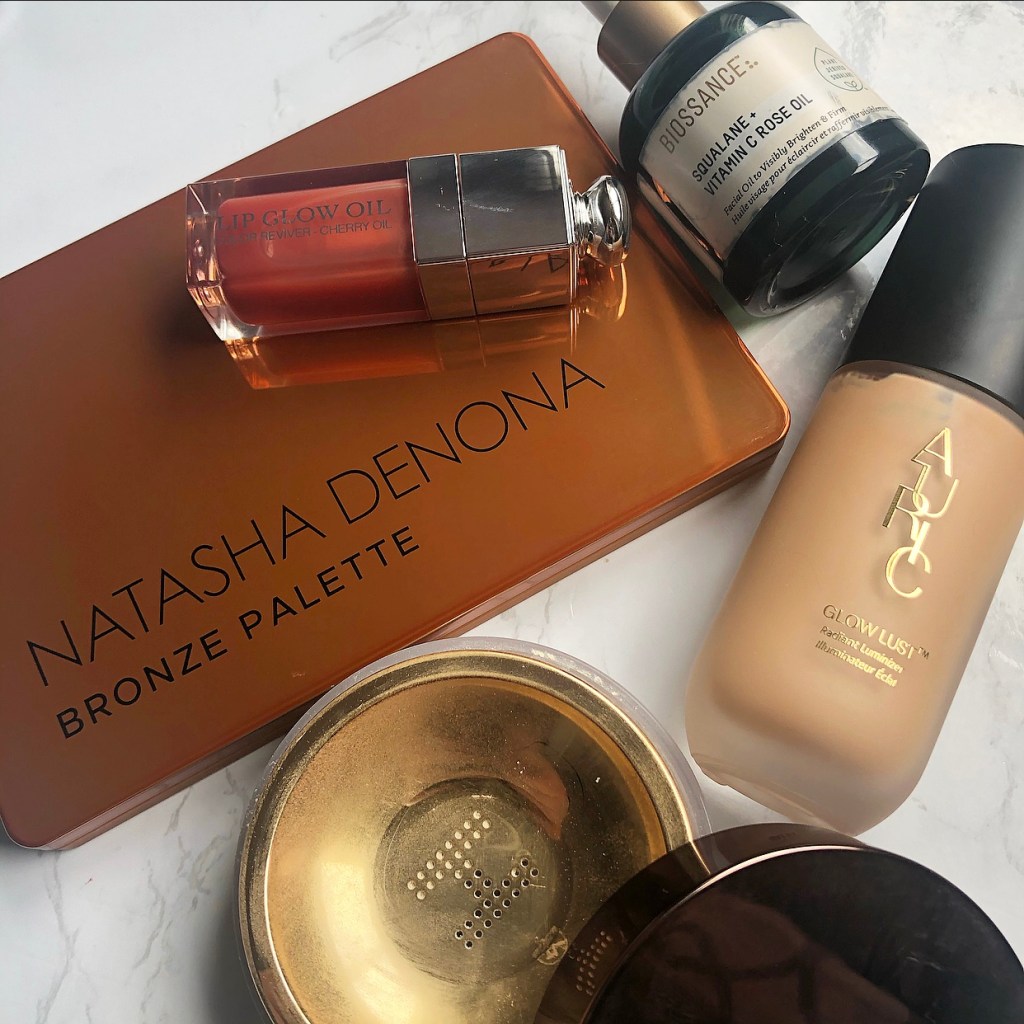 A collection of luxury makeup products including: Natasha Denona Bronze Palette, Dior Lip Glow Oil, Biossance Squalene + Vitamin C Rose Oil, Auric Glow Lust and Hourglass Veil Translucent Setting Powder.